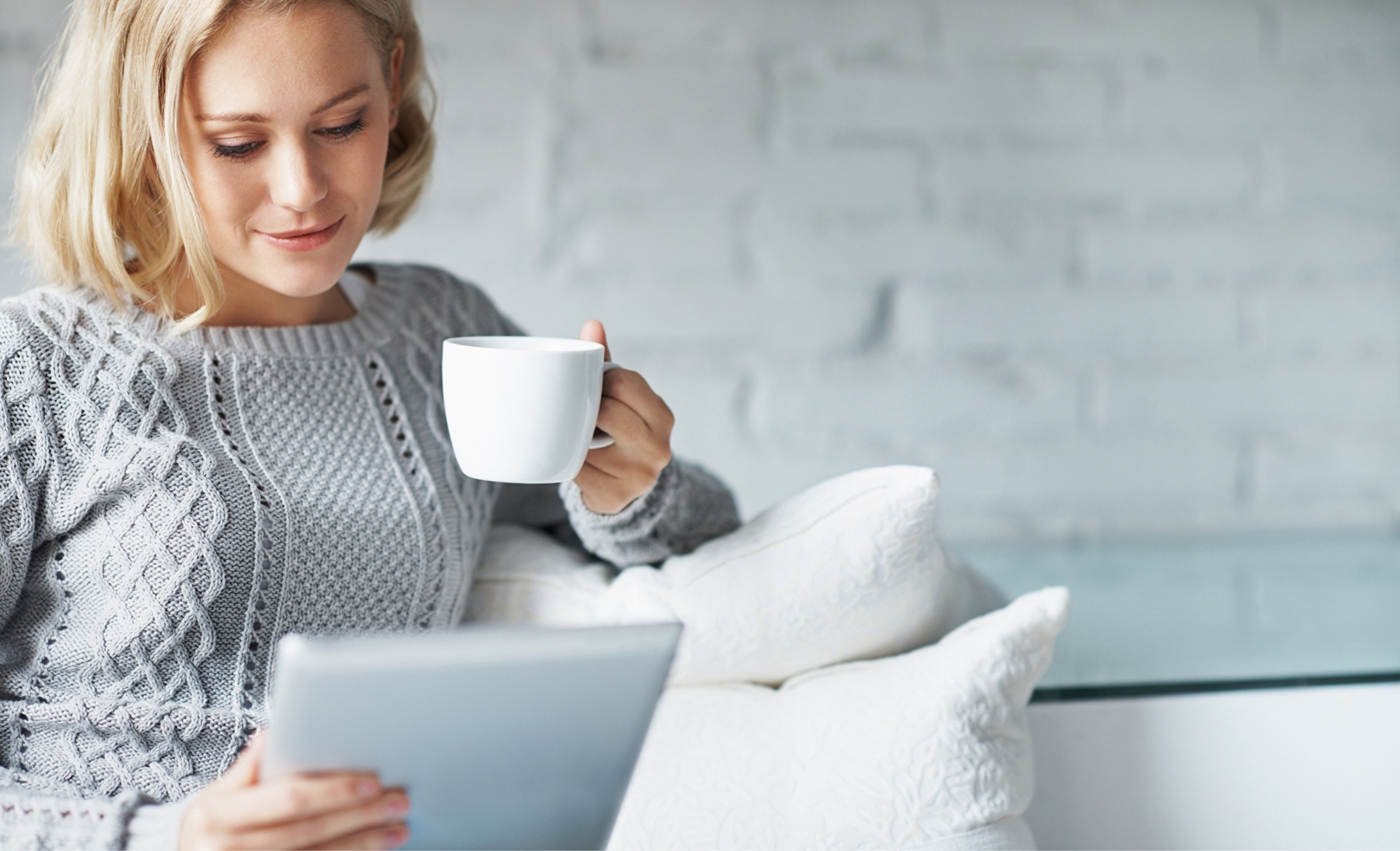 Blonde woman comfortably seated on a sofa while enjoying a cup of coffee and leisurely browsing web content on a tablet.